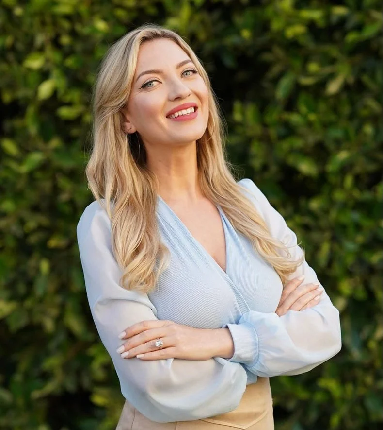 A woman with long blonde hair and fair skin smiling with her arms crossed, standing outdoors in front of green foliage.