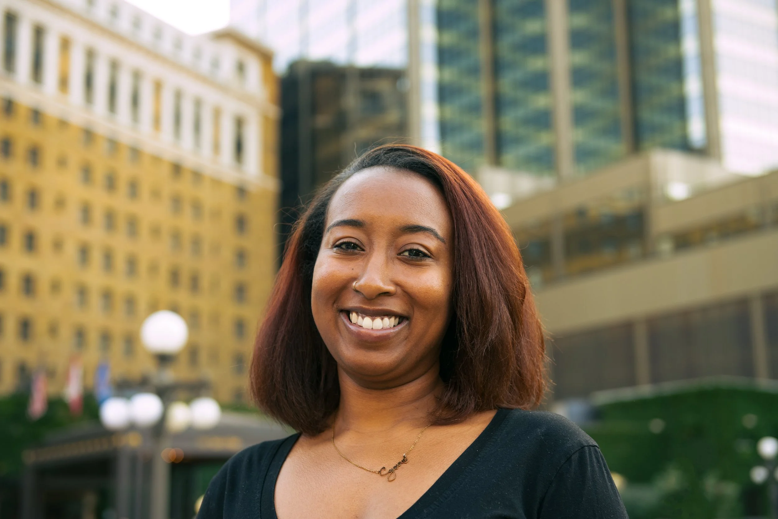Smiling woman with shoulder-length brown hair and a nose piercing, standing outdoors in an urban setting with tall buildings in the background.
