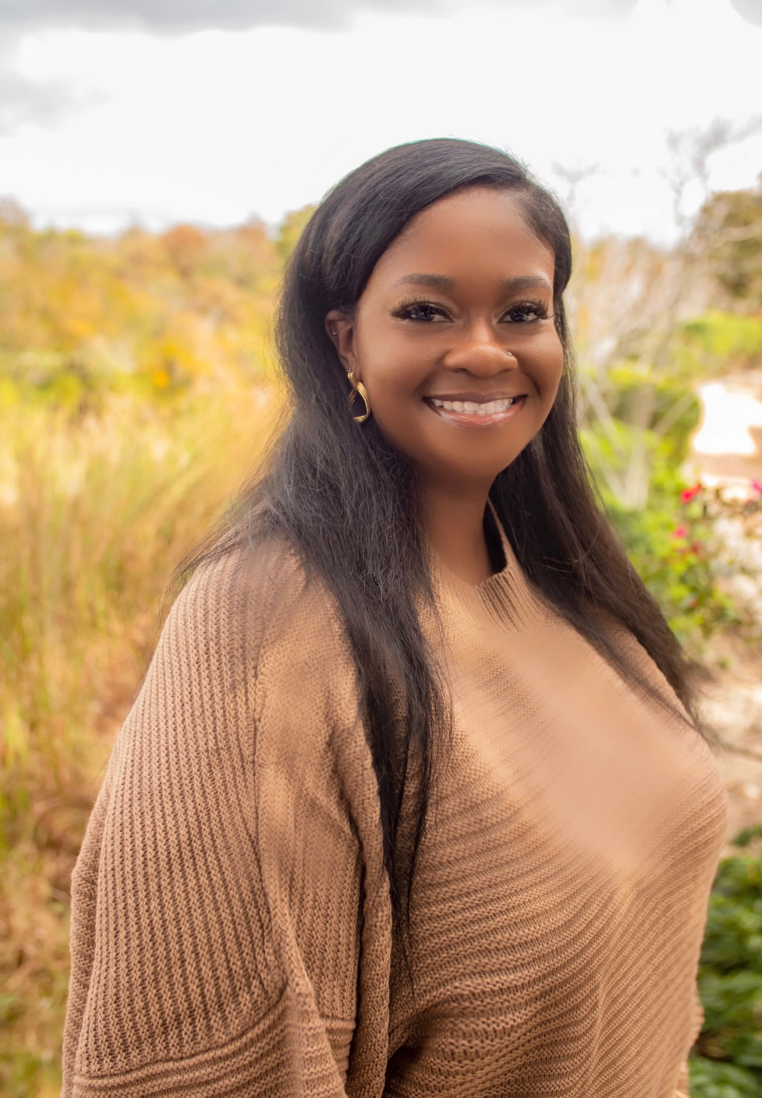 A woman with long dark hair wearing earrings and a tan sweater, smiling outdoors with blurred trees and plants in the background.
