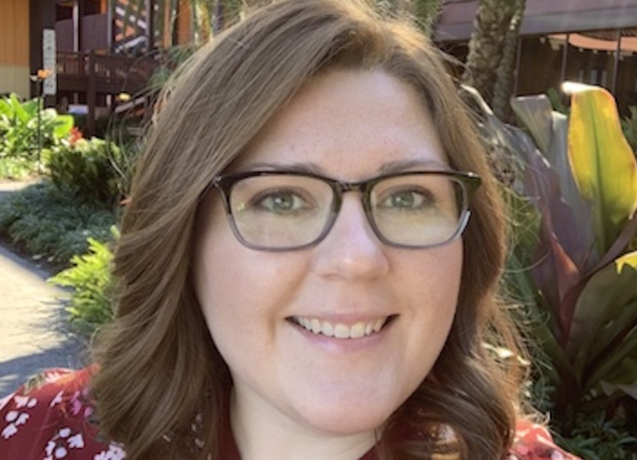A smiling woman with glasses and light brown hair, outdoors in a garden area with plants and trees.