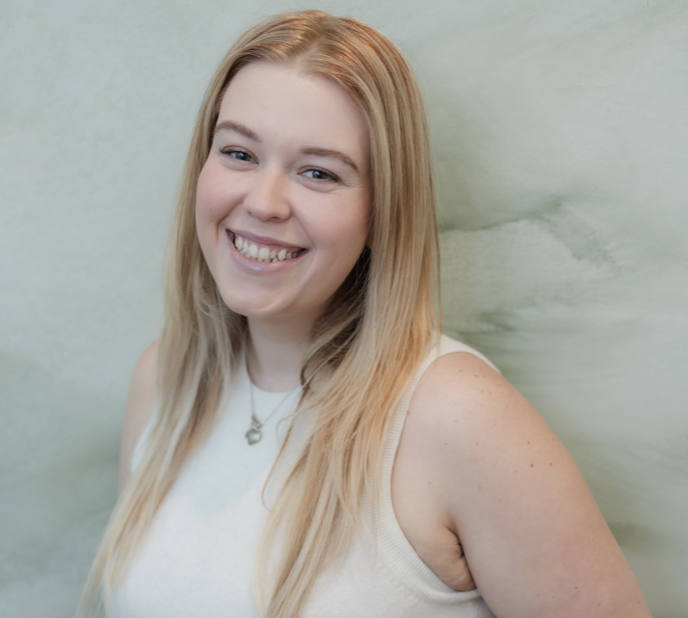 A young woman with long blonde hair smiling with teeth showing, wearing a sleeveless white top and a necklace with a pendant, standing against a light green wall.