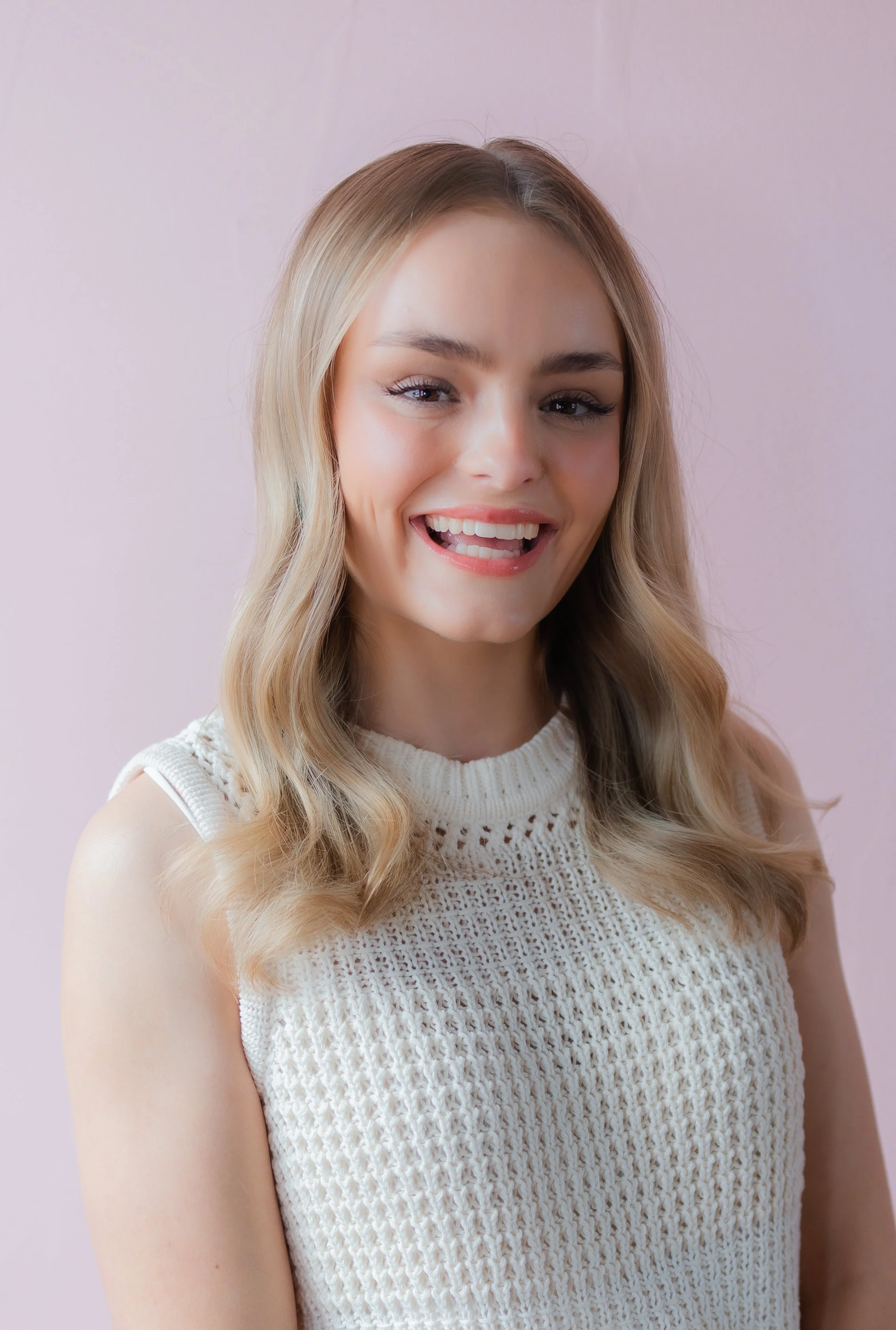 A young woman with blonde hair wearing a sleeveless white knit top, smiling and sticking out her tongue, against a light pink background.