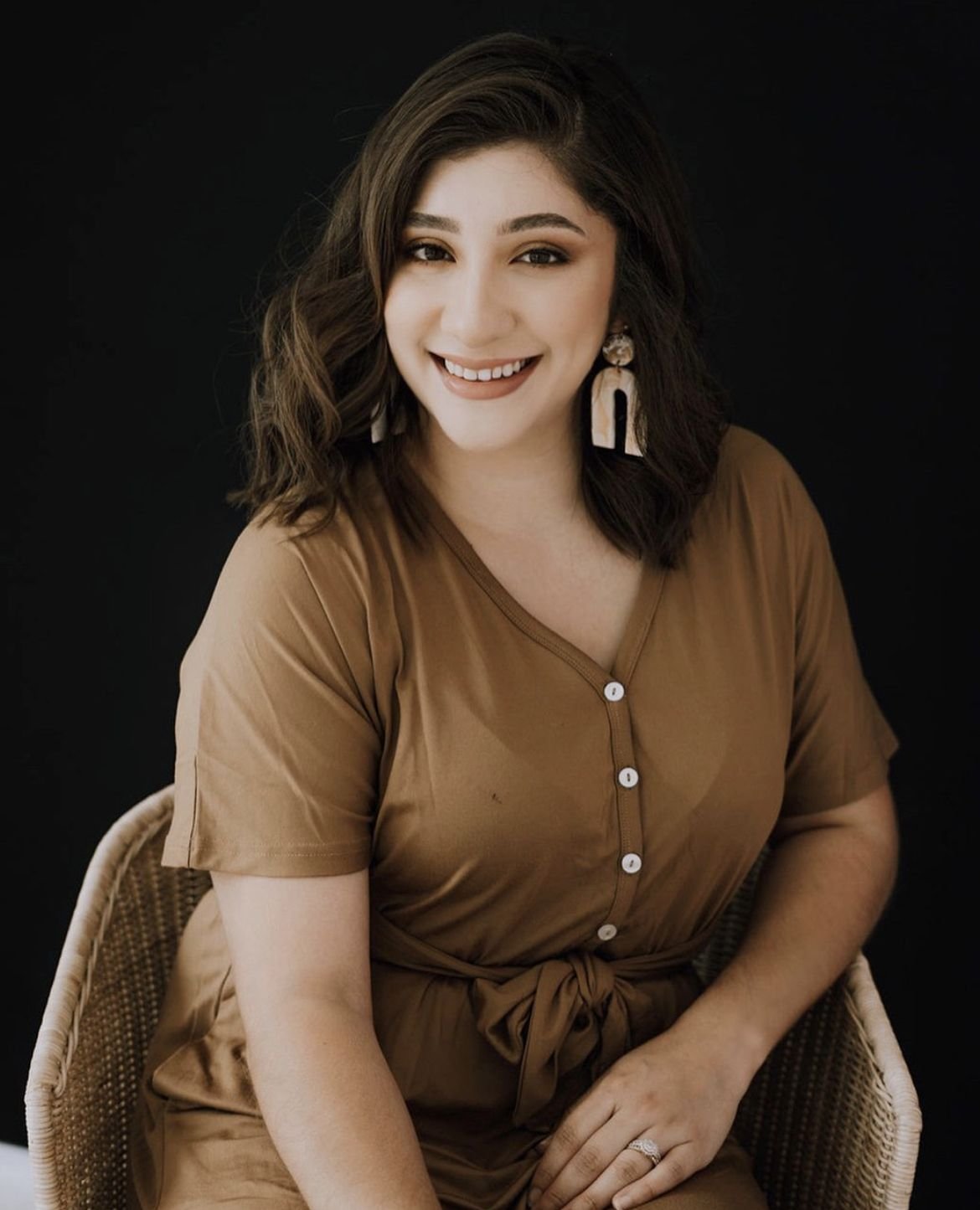 A woman with shoulder-length dark hair and light skin, smiling, wearing a brown button-up dress with a waist tie, statement earrings, and a ring, sitting on a wicker chair against a black background.