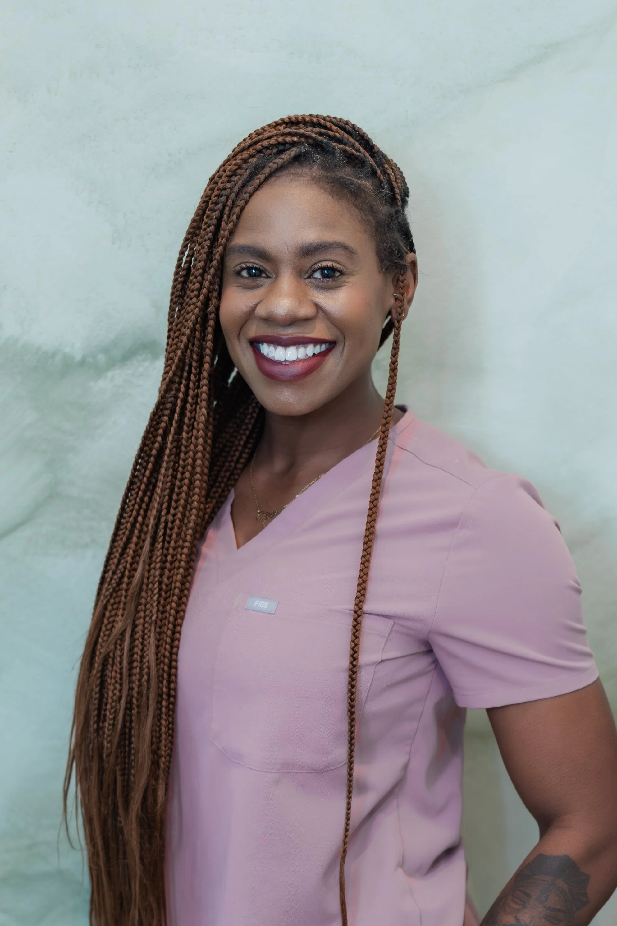 A smiling woman with long braided hair, wearing pink scrubs, standing against a light-colored background.