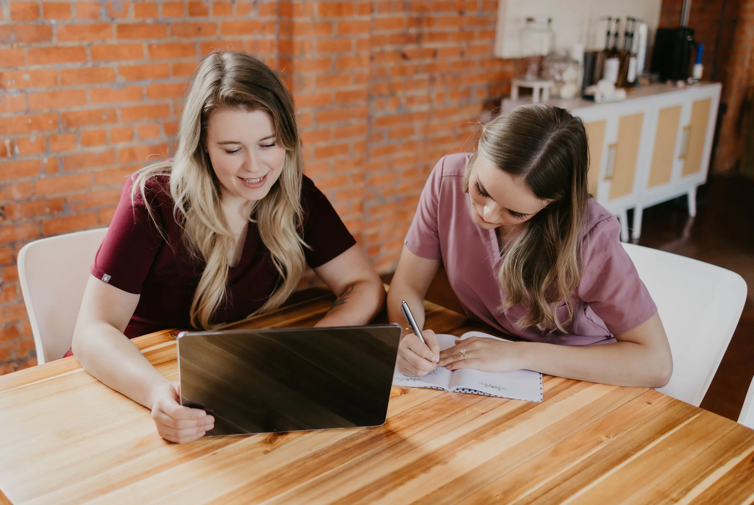 Two women in medical scrubs sitting at a wooden table with a laptop and notebook, smiling and engaged in a discussion in a room with brick walls and a white sideboard in the background.