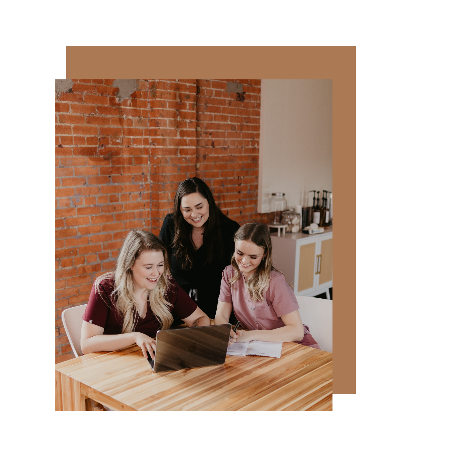 Three women sitting at a wooden table, looking at a laptop, smiling and engaging in a discussion, with a brick wall background.