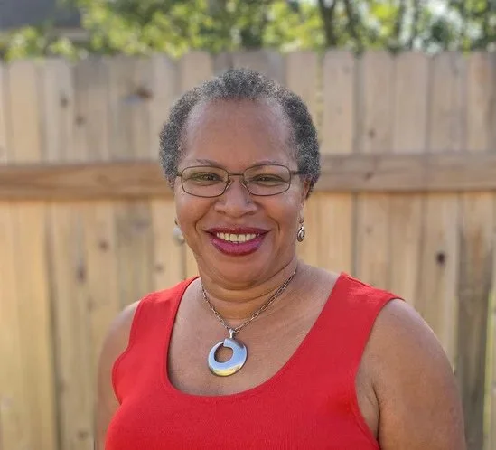 A woman with short curly gray hair and glasses, smiling in front of a wooden fence, wearing a red sleeveless top and a silver necklace with a circular pendant.