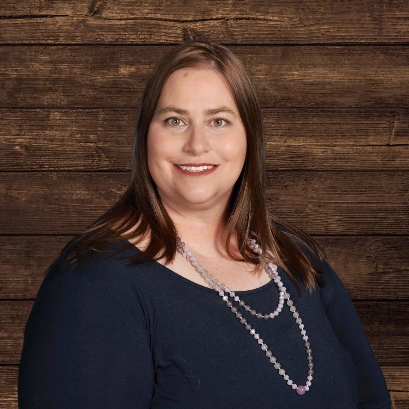 A woman with brown hair and a smile wearing a navy blue top and two pearl necklaces, standing in front of a wooden background.