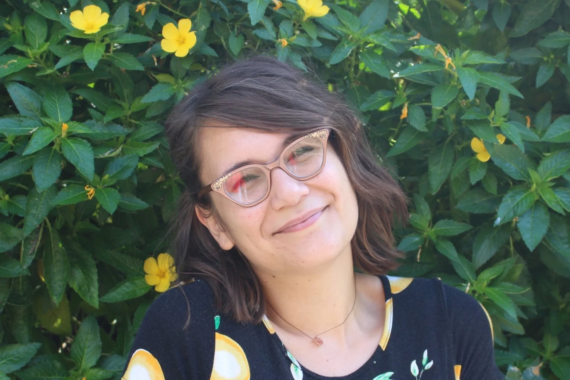 A woman with short brown hair and glasses stands in front of green foliage and yellow flowers, smiling.