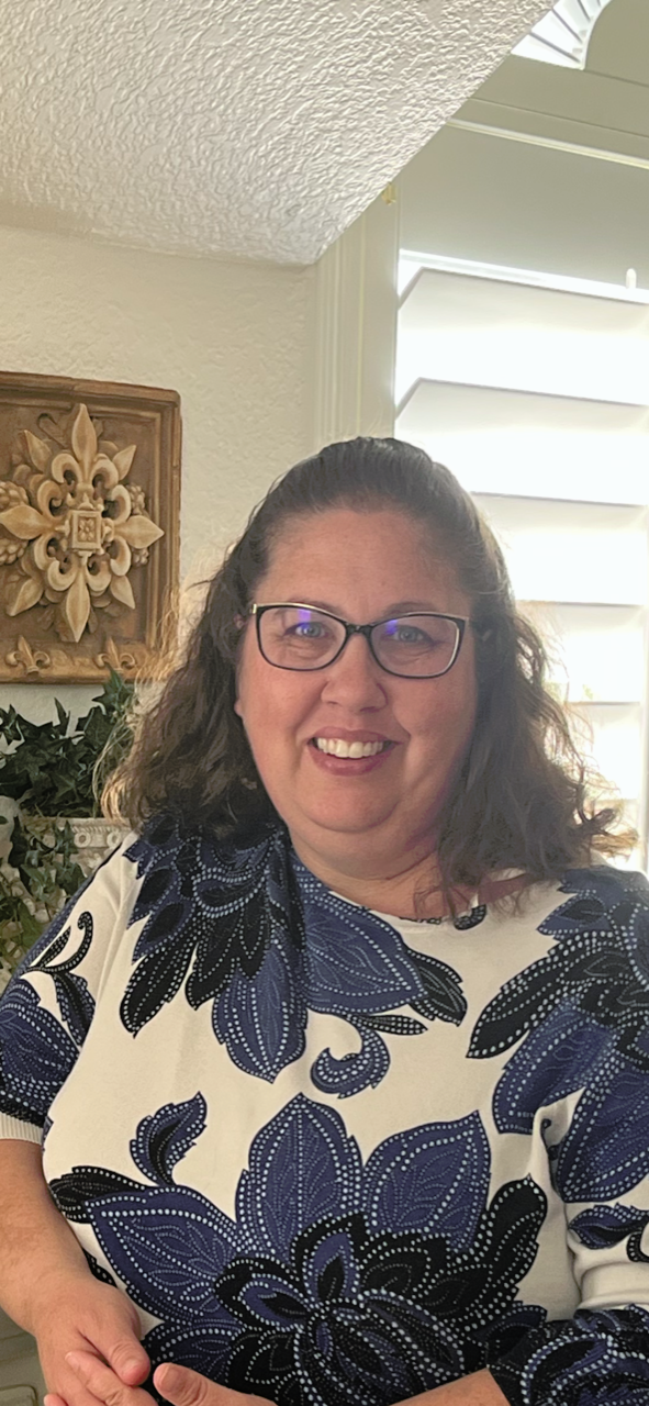 A smiling woman with glasses and curly brown hair, wearing a white shirt with navy blue floral patterns, standing in a room with a decorative wooden wall hanging and greenery behind her.