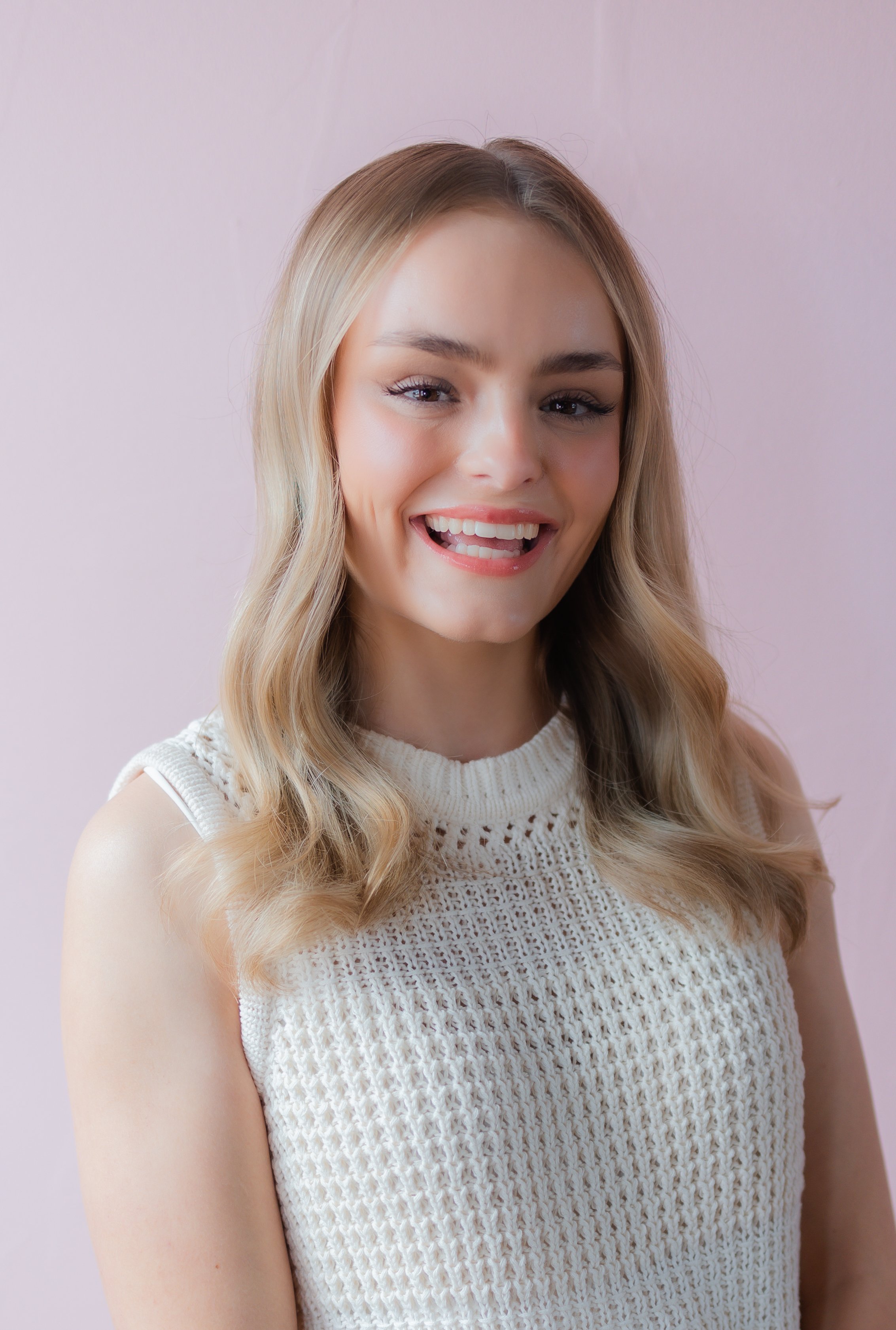 A young woman with blonde hair wearing a sleeveless cream sweater, smiling playfully with her tongue slightly out, against a light pink background.