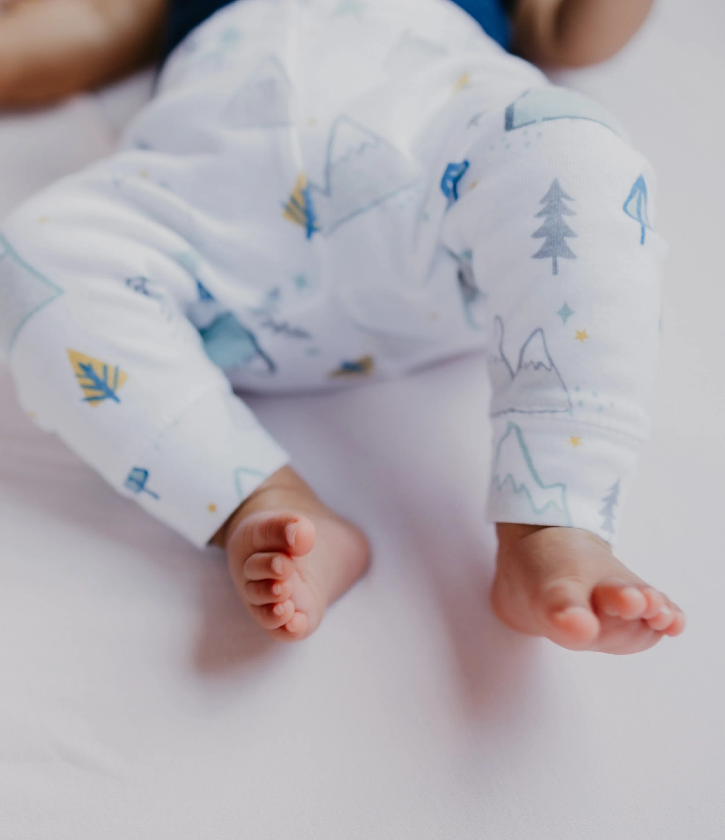 Close-up of a baby lying on their stomach on a white surface, wearing a white outfit with blue, yellow, and gray mountain and tree patterns.