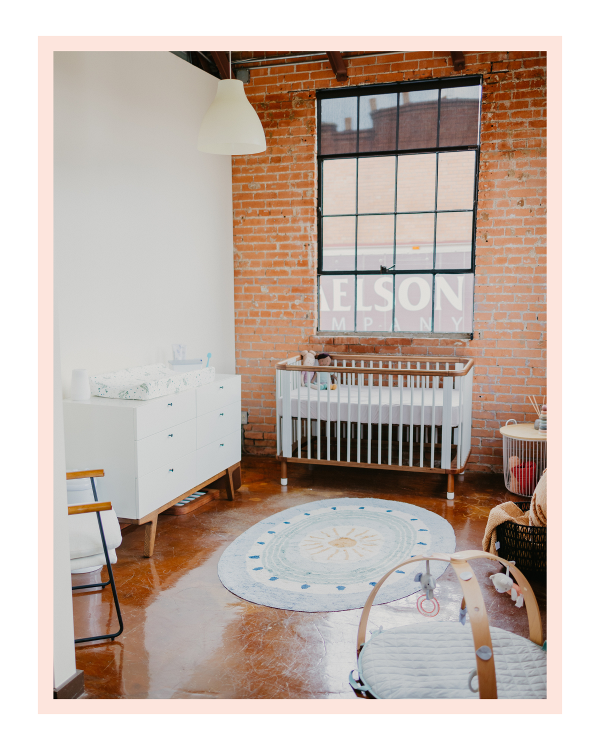 Nursery room with exposed brick wall, large window, white crib with stuffed animals, white dresser, circular rug with sun design, and a baby activity mat.