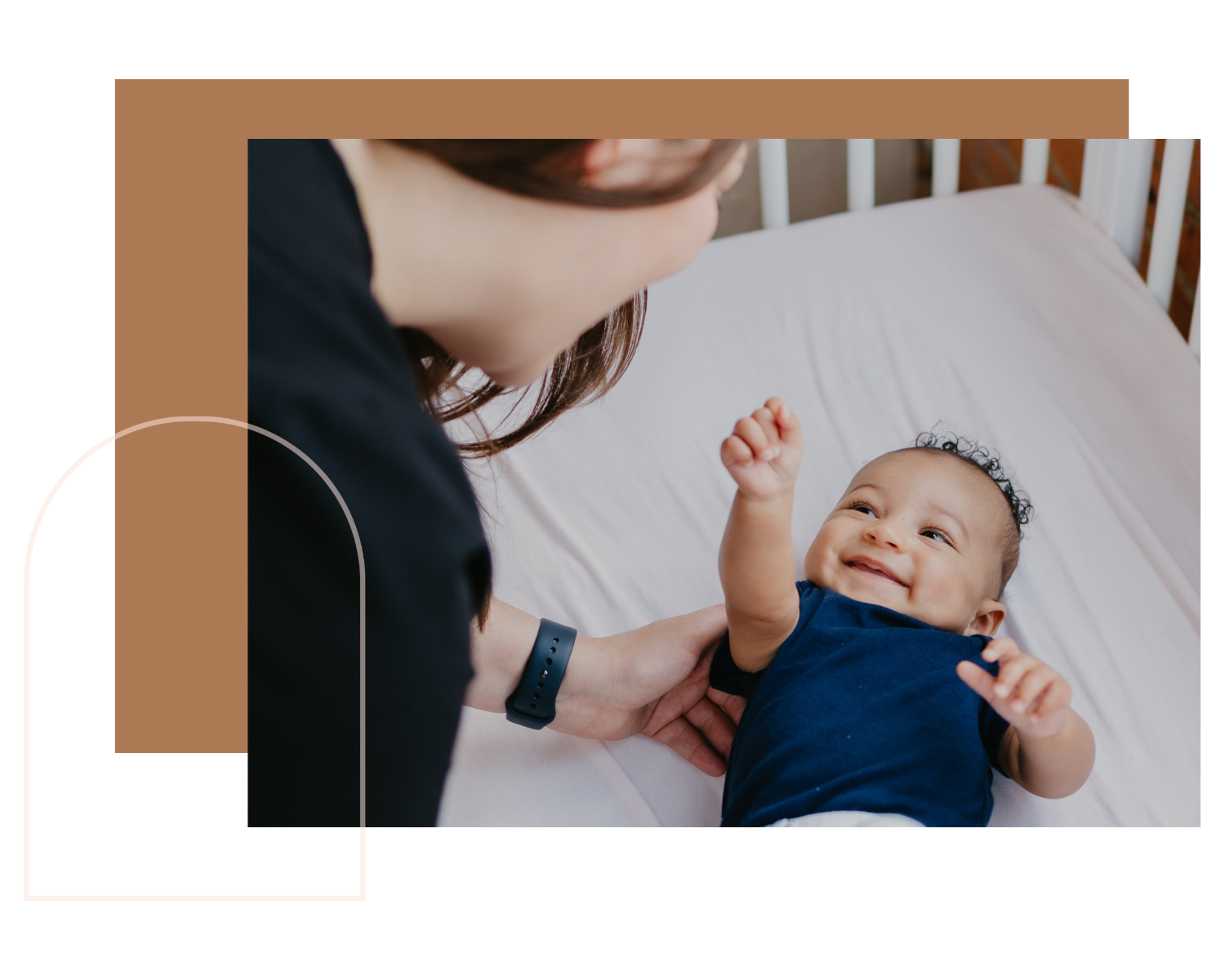 A woman with brown hair and a black top lying on a bed, smiling and looking at a happy baby lying on his back on a white sheet. The baby is wearing a navy blue shirt and is reaching upward.