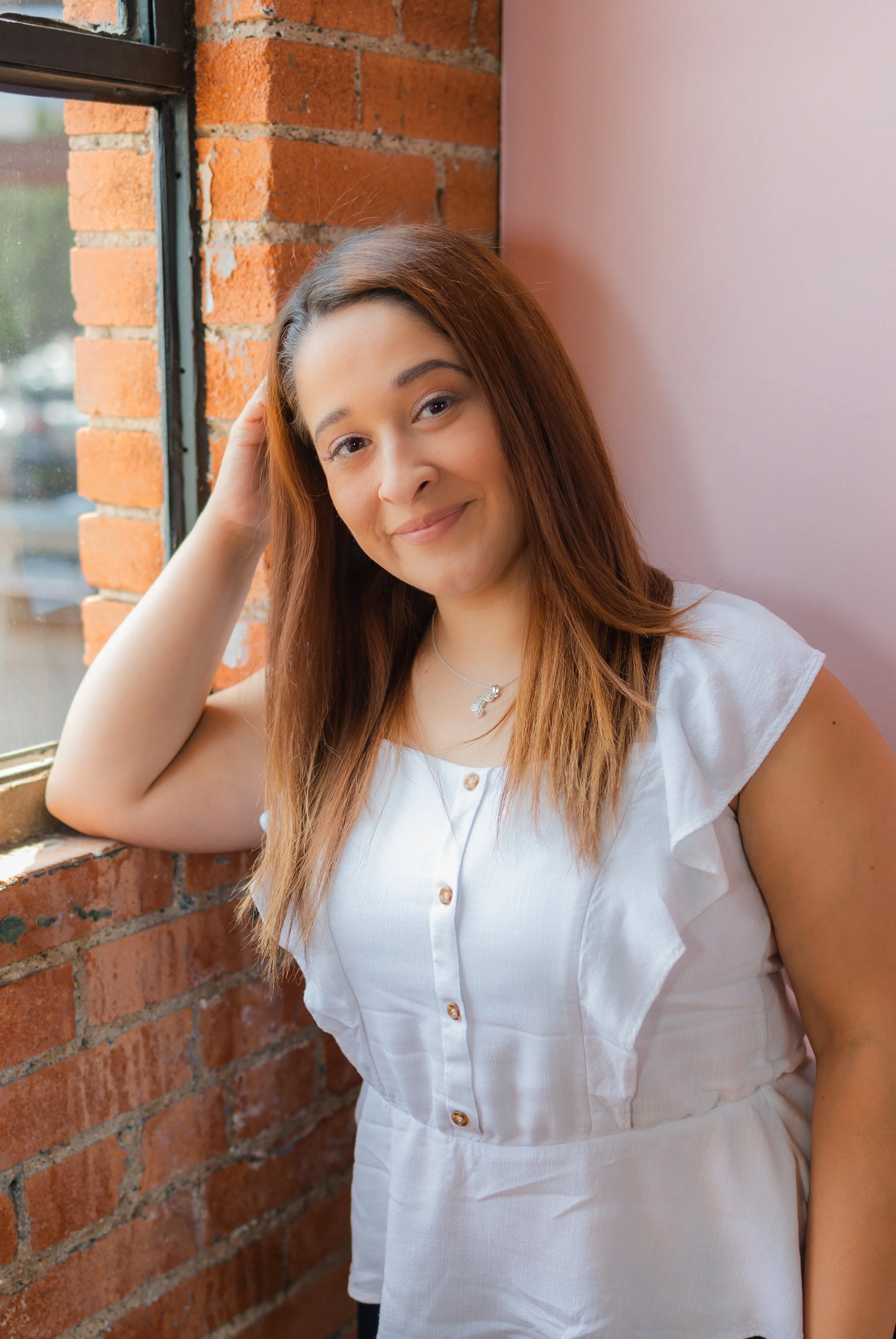 A woman with long brown hair leaning against a brick wall next to a window, smiling at the camera, wearing a white blouse and a silver necklace.
