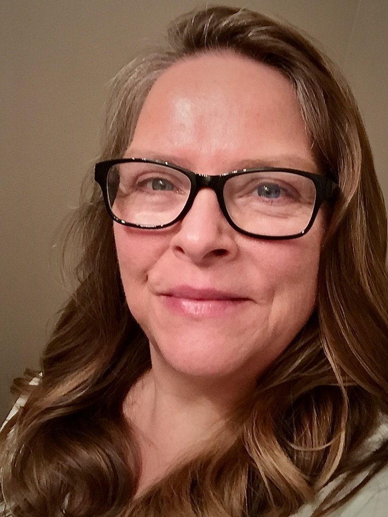 A woman with long, wavy brown hair wearing black glasses, smiling slightly, against a plain beige background.