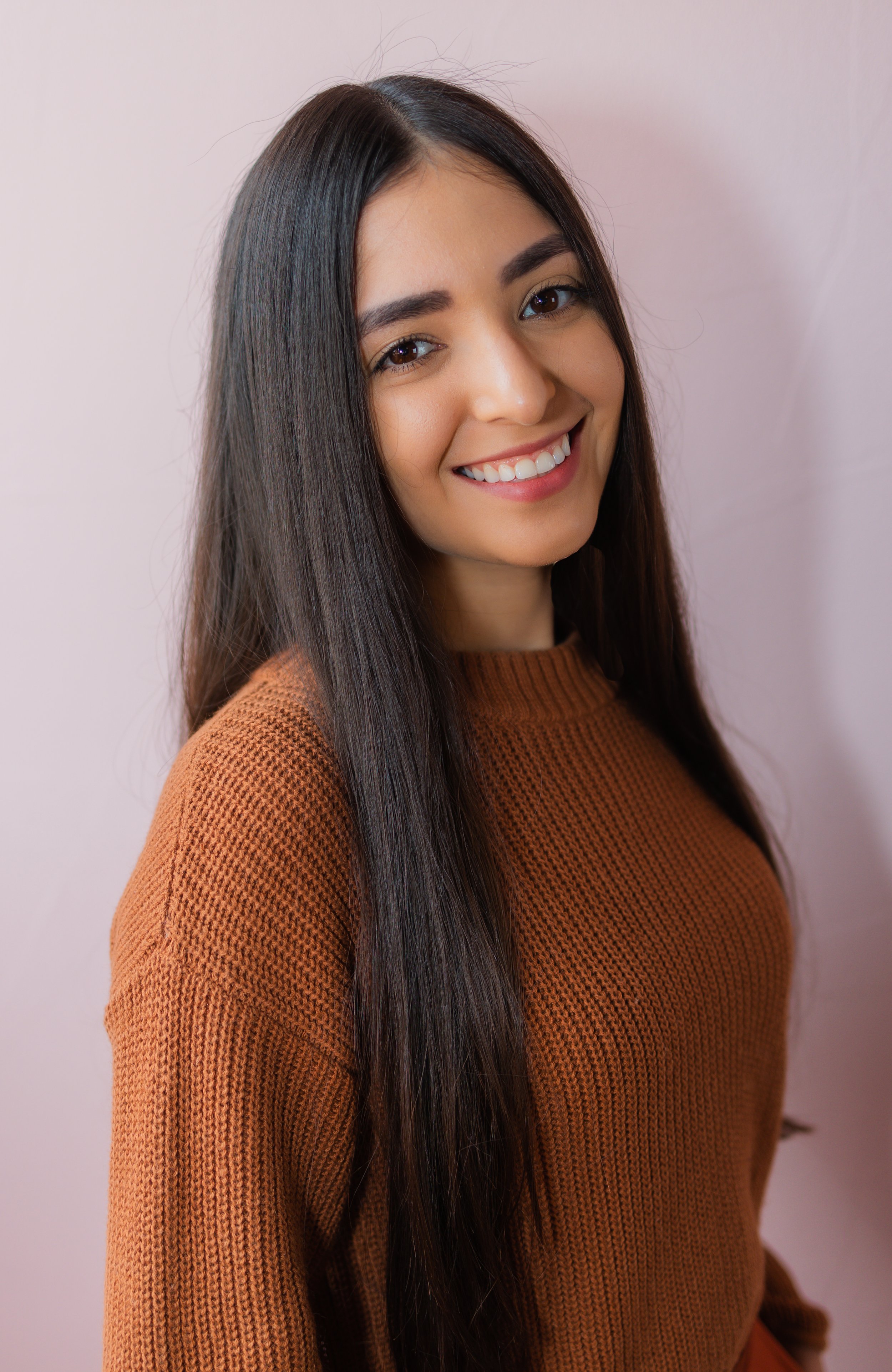 A young woman with long dark hair smiling, wearing a brown sweater, against a light pink background.