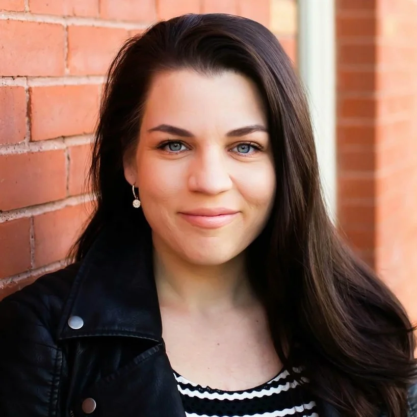 A woman with long dark hair, blue eyes, wearing earrings and a black leather jacket, smiling in front of a brick wall.