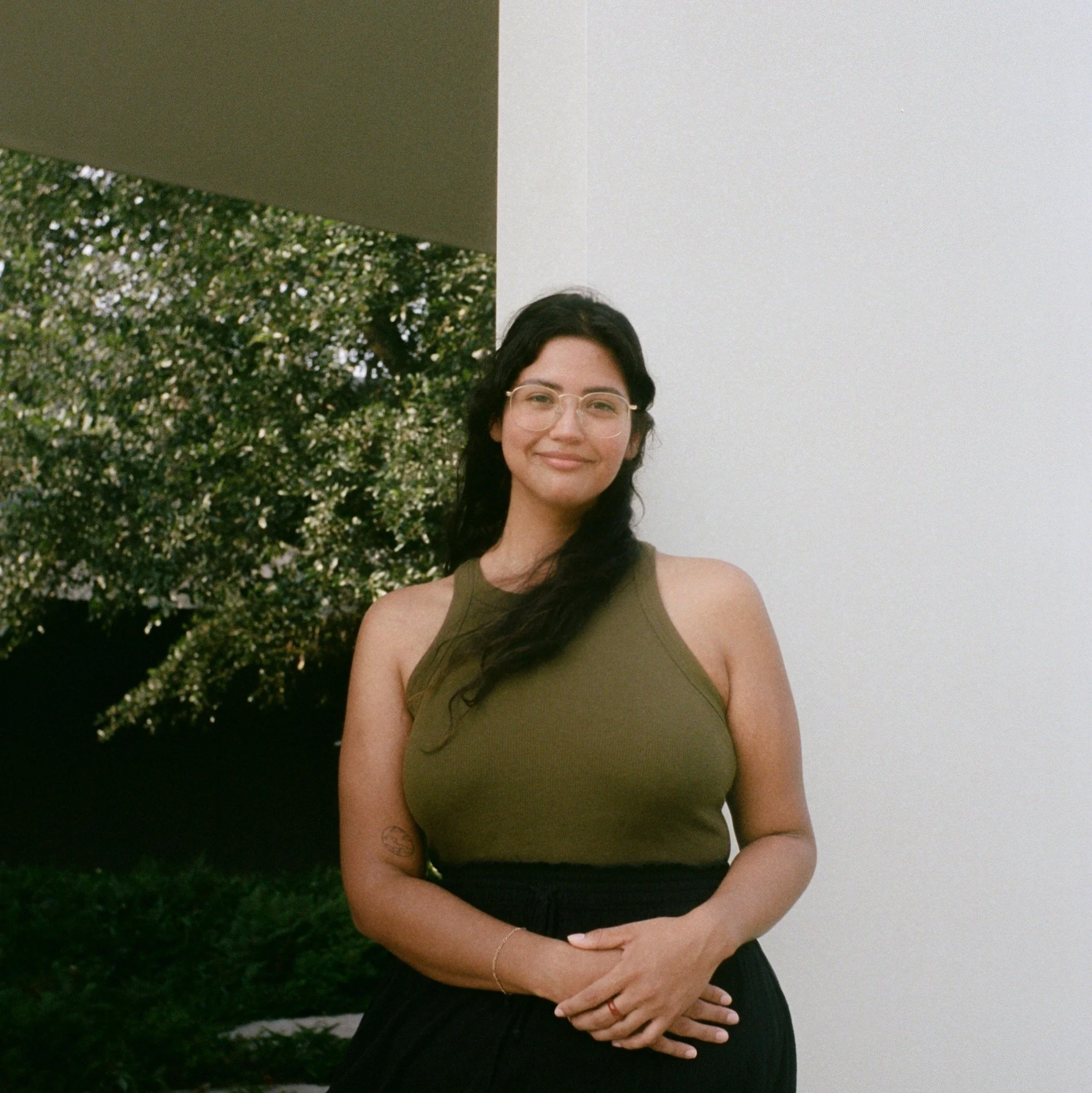 A woman with long dark hair, glasses, a sleeveless olive green top, and black pants standing outdoors near a modern building with a white wall and trees in the background.