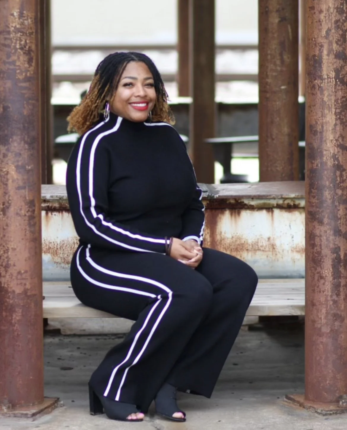 A woman with curly hair, wearing a black tracksuit with white stripes, sitting on a wooden bench under a rusted metal structure, smiling at the camera.