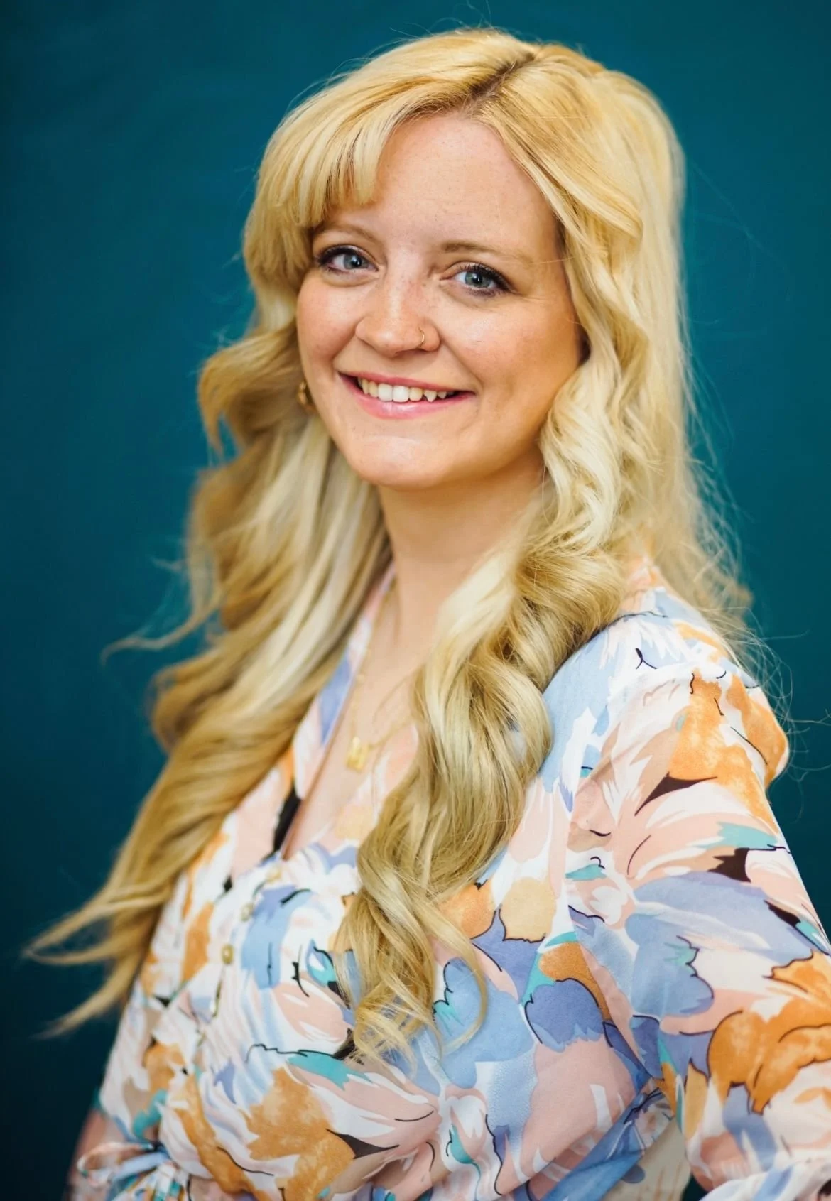 A woman with long, wavy blonde hair, wearing a floral blouse in shades of blue, orange, and beige, smiling against a teal background.