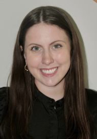 A woman with long brown hair, wearing a black top, smiling at the camera.