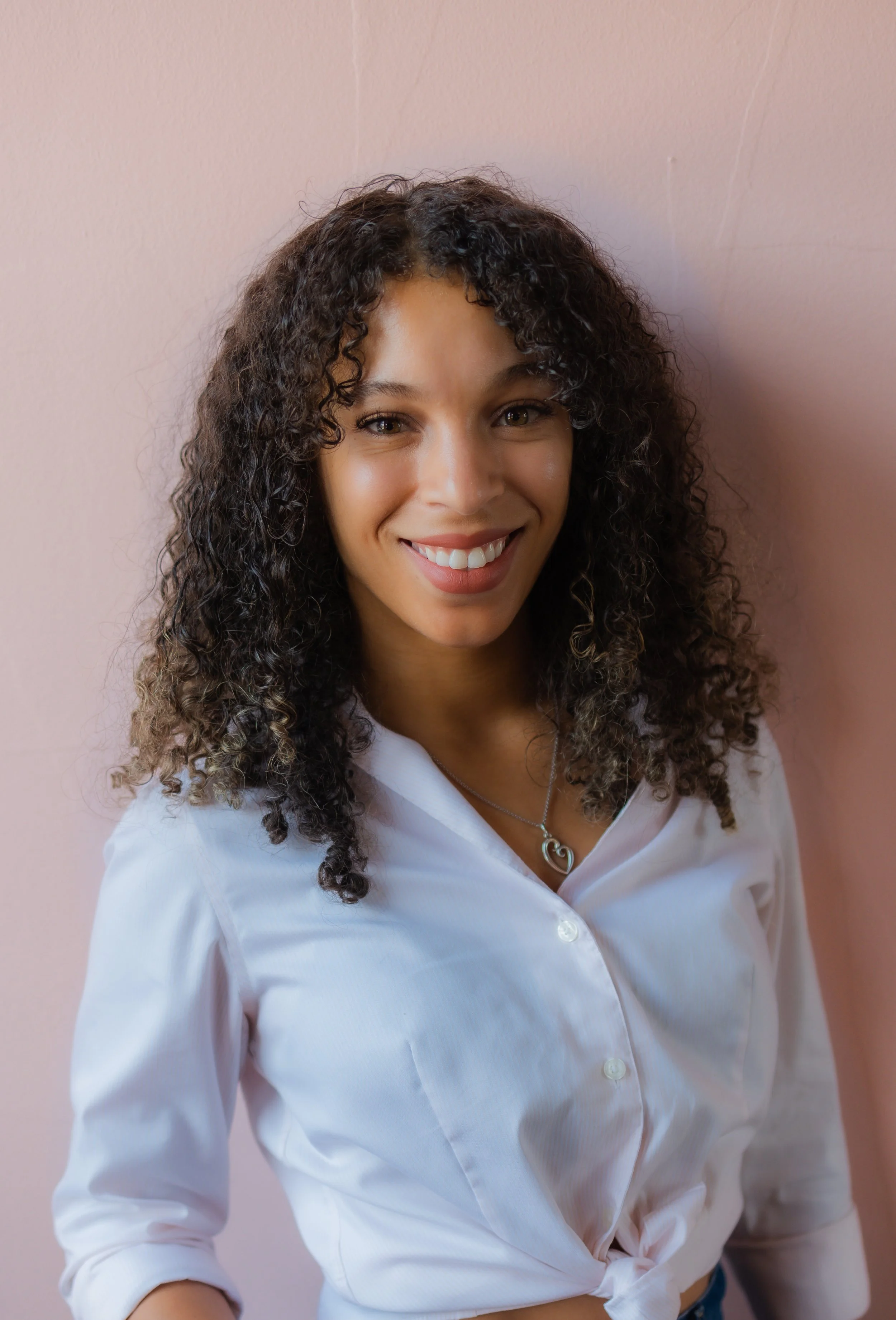 A young woman with curly brown hair, smiling, wearing a white button-up shirt and a heart-shaped necklace, standing against a light pink wall.