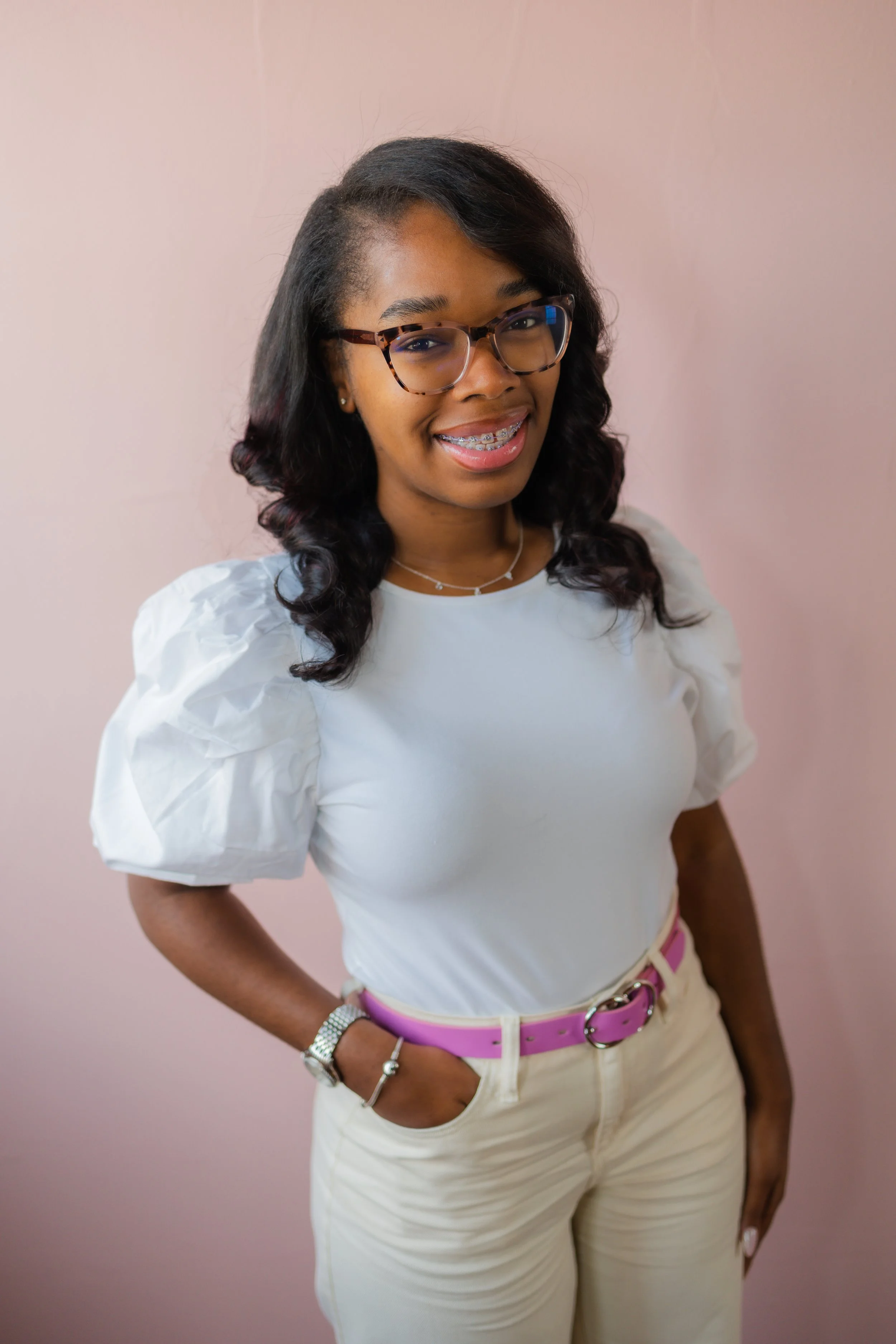 A young woman with glasses, braces, and wavy hair, smiling and standing with her hand in her pocket against a pink background, wearing a white top with puff sleeves and beige pants with a pink belt.