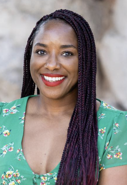 A woman with long purple braids smiling outdoors, wearing a green floral dress.