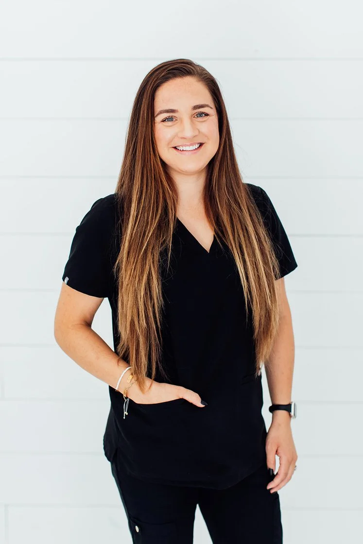 A young woman with long brown hair smiling at the camera, wearing a black medical scrub top and a watch, standing against a white paneled background.
