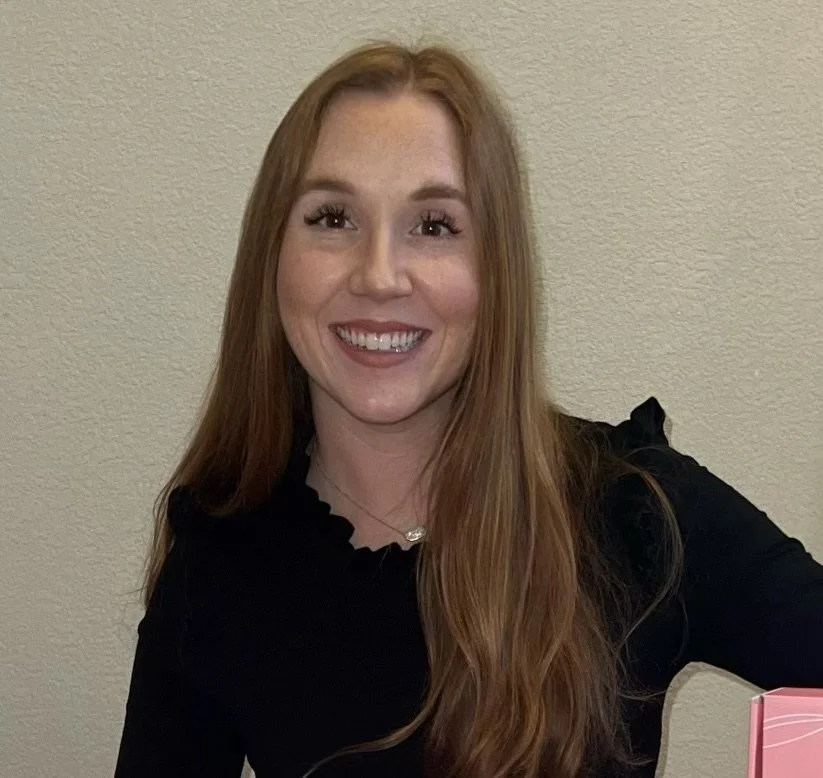 A woman with long red hair, smiling, wearing a black top, sitting in front of a beige wall.