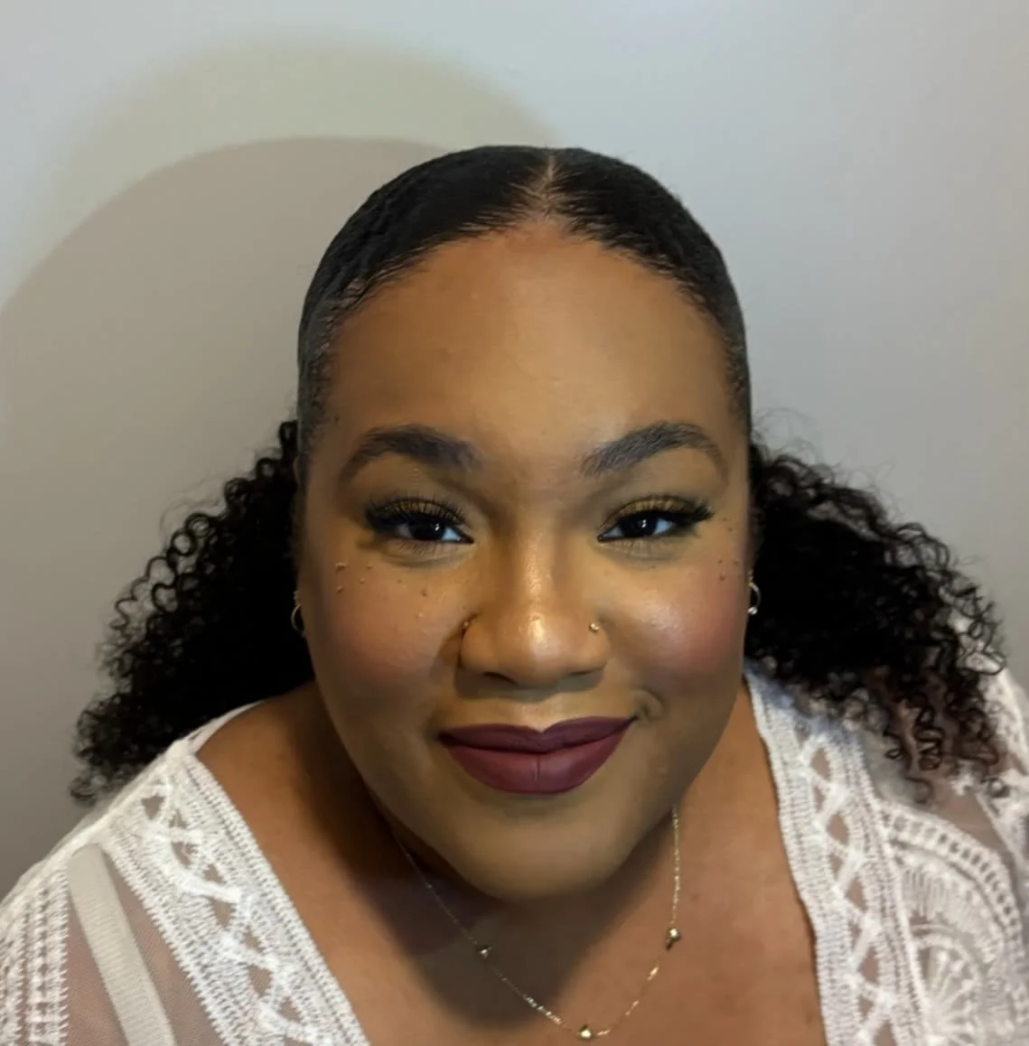 A woman with dark curly hair, wearing makeup and earrings, looking at the camera with a slight smile, dressed in a white lace top