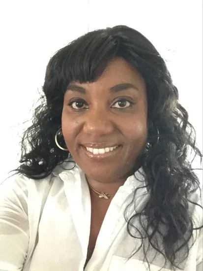 A woman with shoulder-length curly black hair, wearing hoop earrings and a white button-up shirt, smiling at the camera.