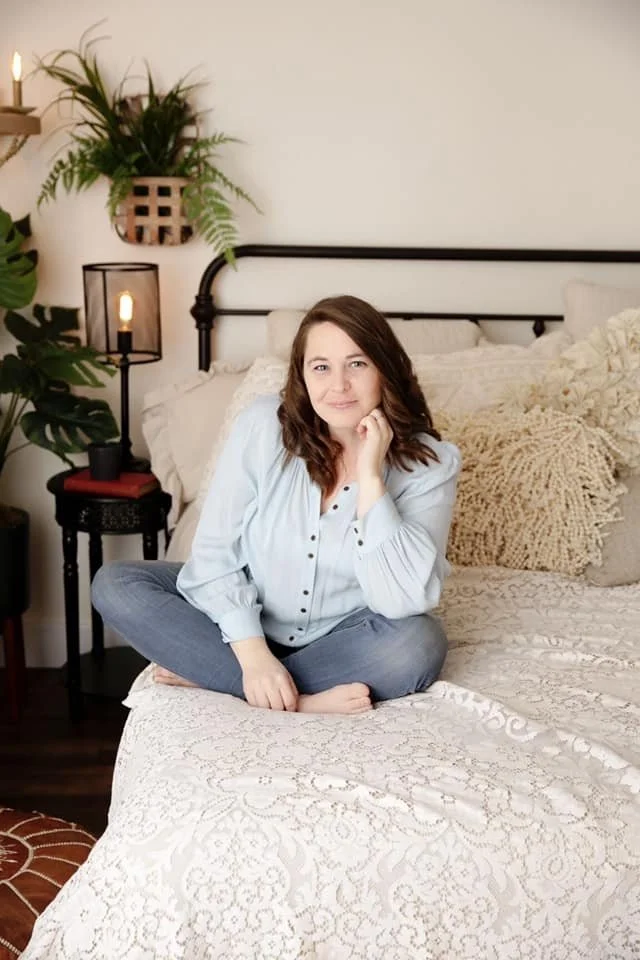 A woman sitting cross-legged on a bed in a bedroom, smiling and resting her chin on her hand, with plants and bedside lamps in the background.