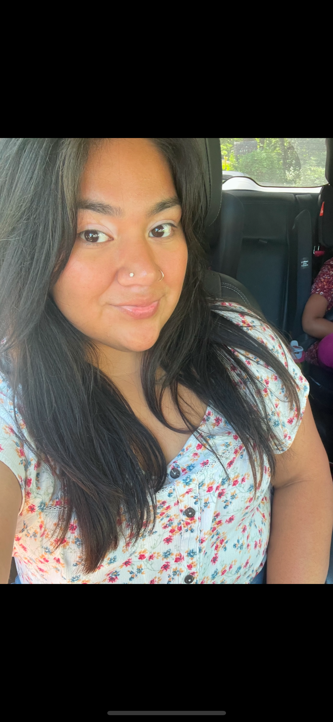 A woman with long dark hair, a nose piercing, and a light-colored floral top taking a selfie in a car, with a child in the backseat.