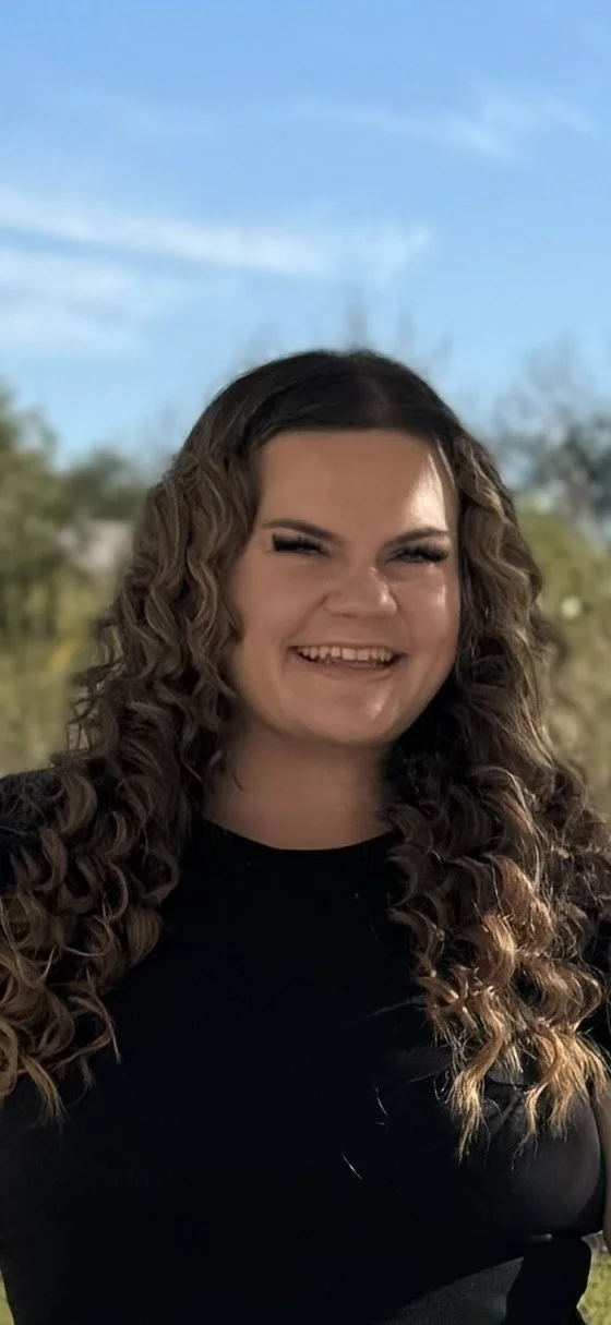 A young woman with long, curly brown hair smiling outdoors against a blue sky and trees.