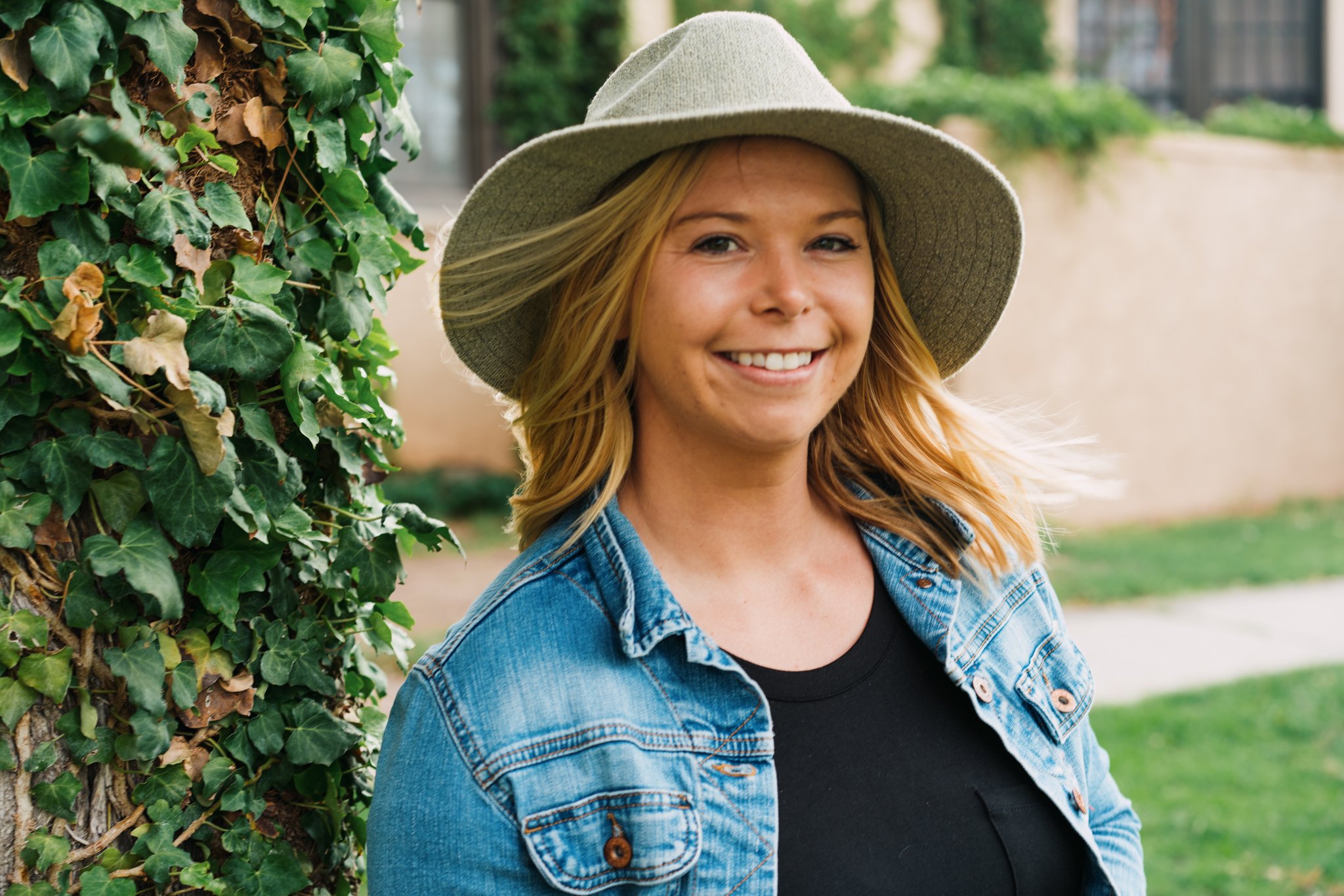 A smiling woman with blonde hair wearing a wide-brimmed hat and a denim jacket, standing outdoors near green ivy.