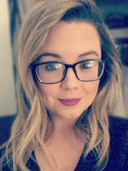 A woman with long blonde hair and glasses, smiling and looking down.