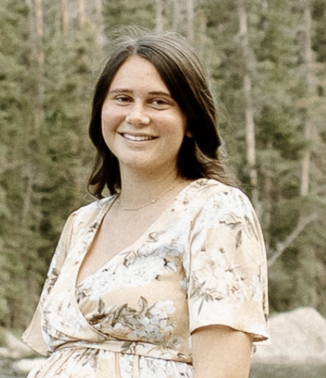 A smiling young woman with shoulder-length dark hair standing outdoors in a wooded area.
