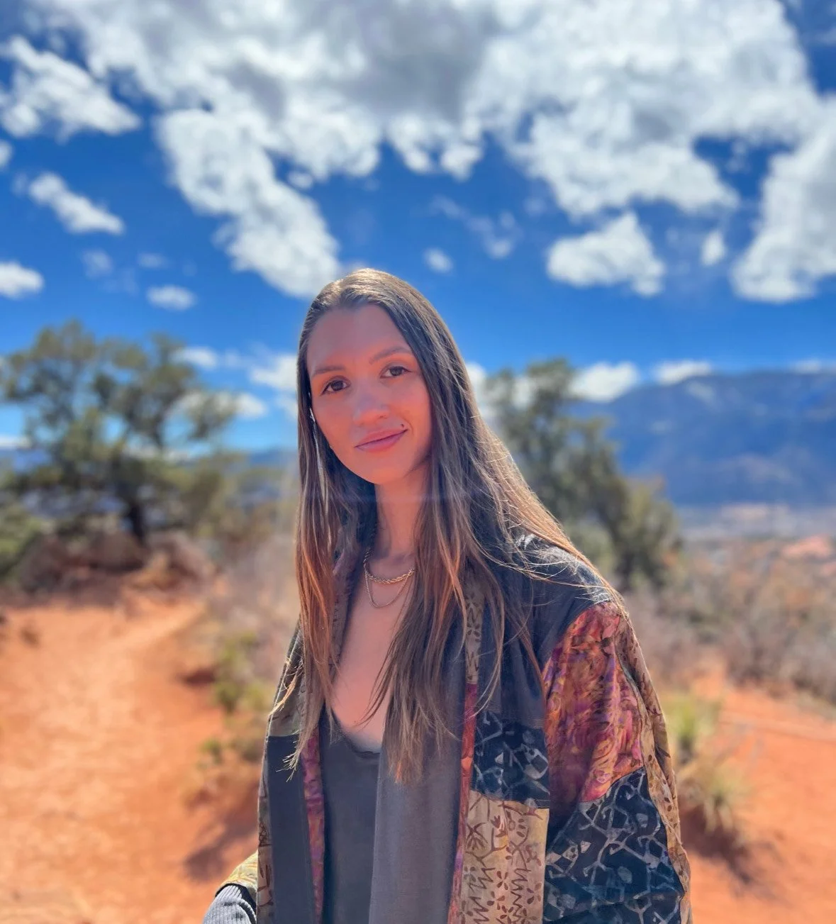 A young woman with long brown hair and a necklace standing outdoors in a desert landscape under a partly cloudy blue sky.