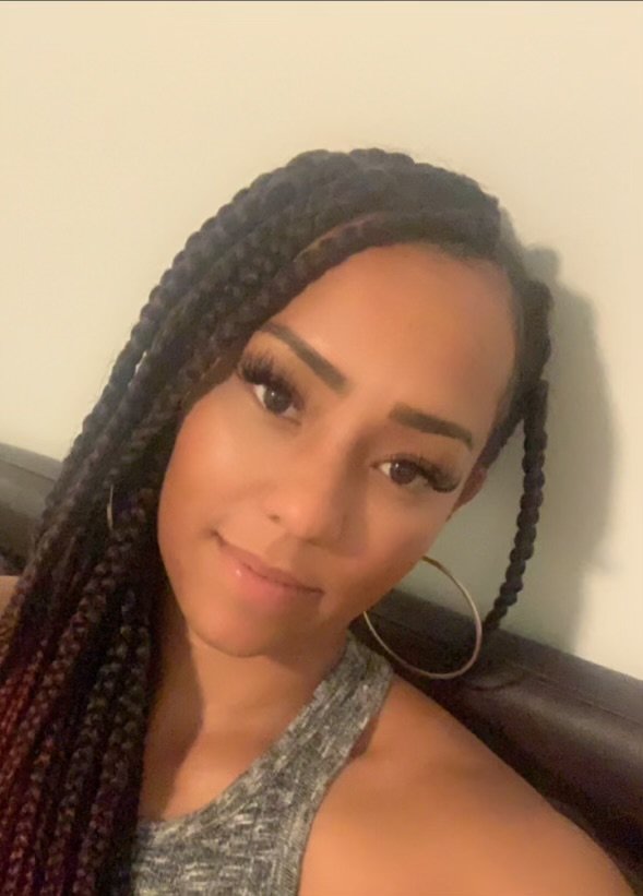 A woman with long braided hair, wearing large hoop earrings and a gray sleeveless top, smiling at the camera indoors.