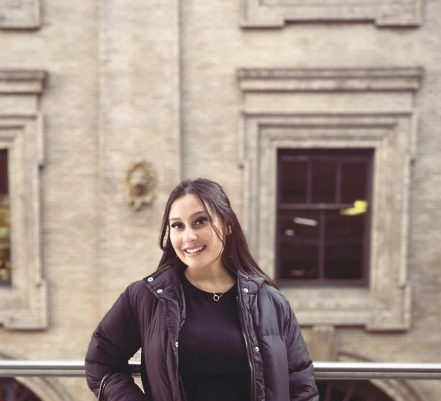 A young woman standing outside in front of an old brick building, smiling at the camera, wearing a black jacket and black top.