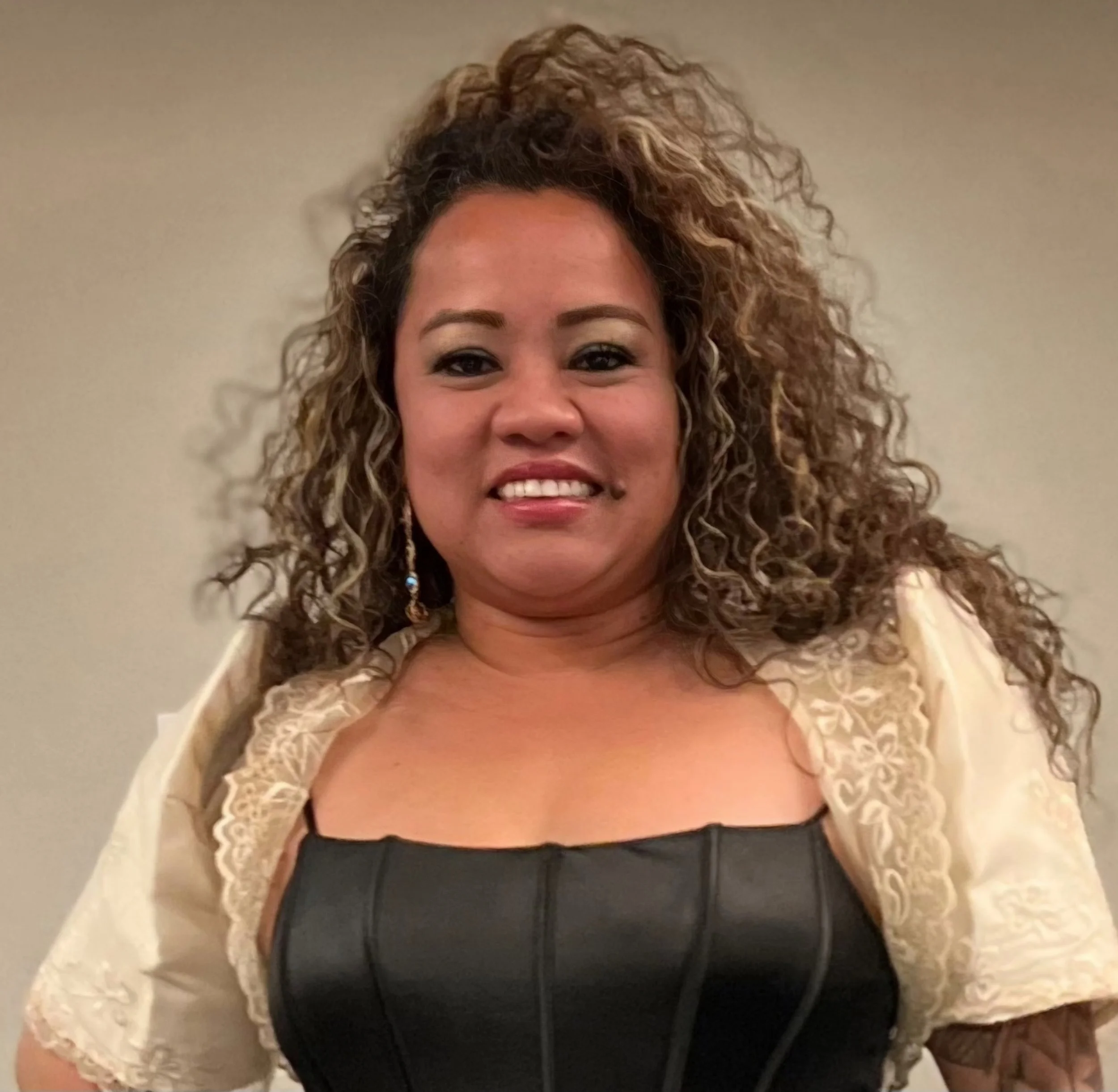 A woman with curly hair smiling, wearing earrings, a black leather top, and a cream-colored lace jacket.