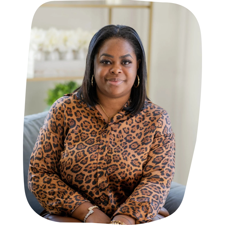 A woman with medium-length black hair wearing a leopard print blouse, gold hoop earrings, and a gold necklace, sitting indoors with a blurred background including some plants.