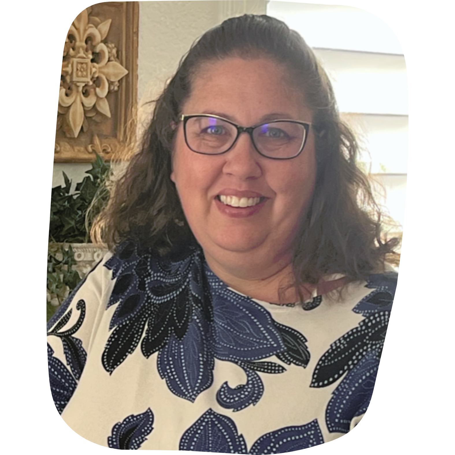 A woman with glasses and shoulder-length dark hair, smiling in front of a beige wall with a decorative wooden wall hanging and green plants.