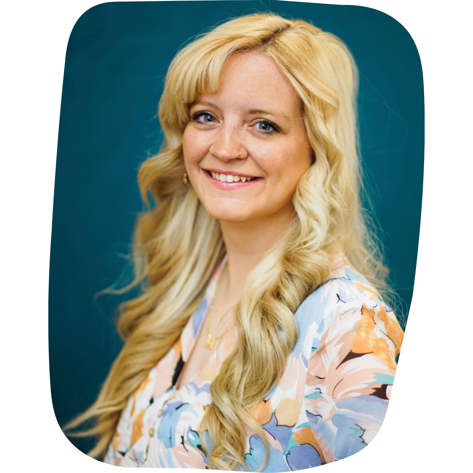 A woman with long blonde wavy hair, blue eyes, and fair skin, smiling and wearing a colorful floral blouse, against a teal background.