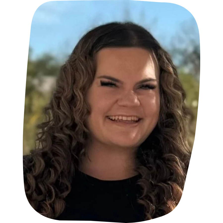 A woman with curly brown hair smiling outdoors with trees and blue sky in the background.