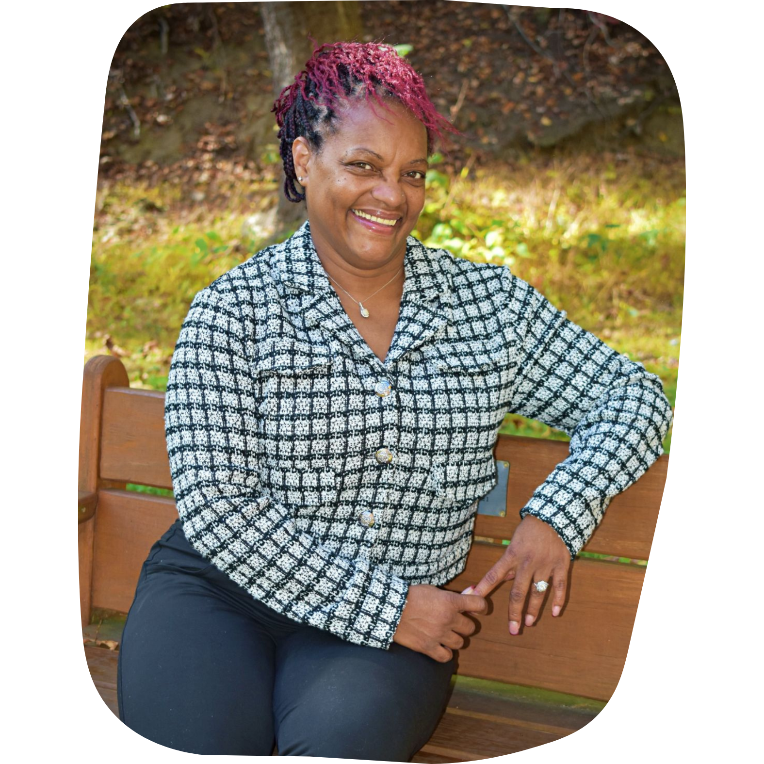 A smiling woman with short, pink and purple braided hair, wearing a black and white checkered jacket with gold buttons, sitting on a wooden park bench outdoors with a background of trees and greenery.