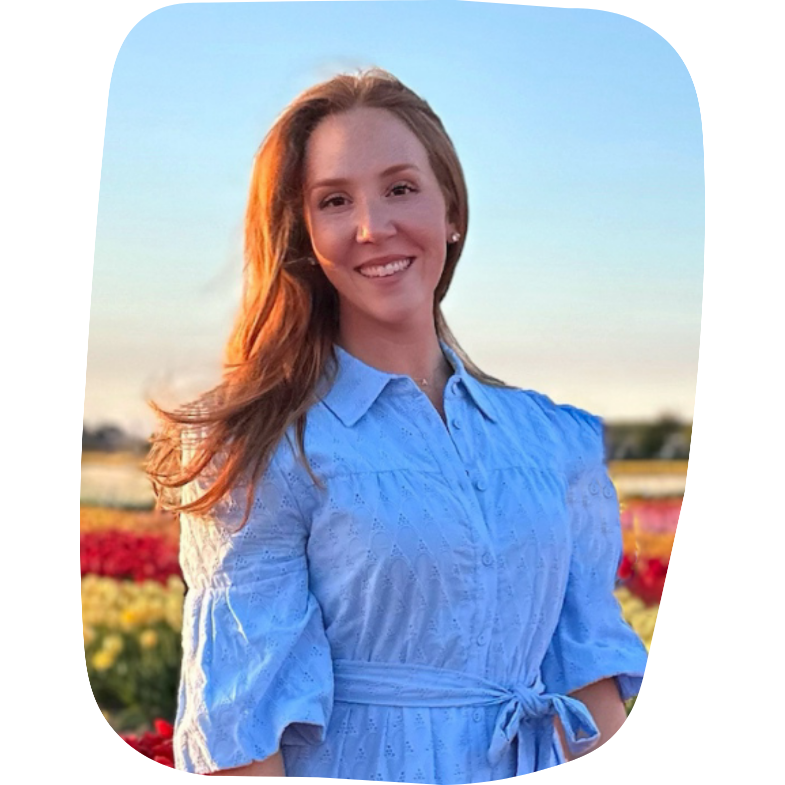 A woman with long red hair smiling outdoors in a field of colorful flowers during sunset, wearing a light blue dress with puffed sleeves and a waist tie.