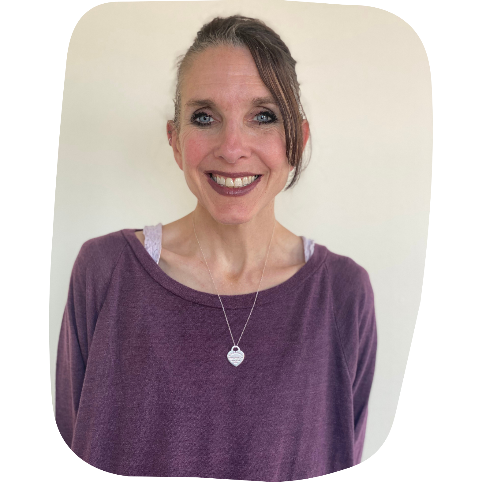 A woman smiling, wearing a purple long-sleeve shirt, with a heart-shaped necklace, standing against a plain white wall.